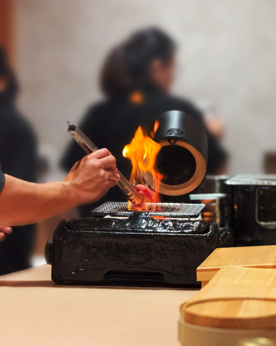 A chef carefully grills premium ingredients over open flame in an authentic omakase setting, showcasing precision, tradition, and refined craftsmanship.