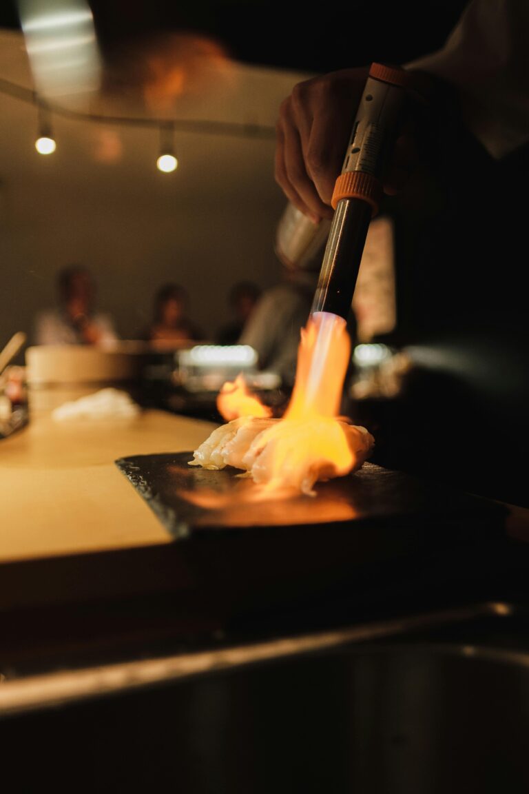 A sushi chef lightly searing a piece of premium fish with a handheld burner, creating a delicate smoky aroma and showcasing refined omakase technique.