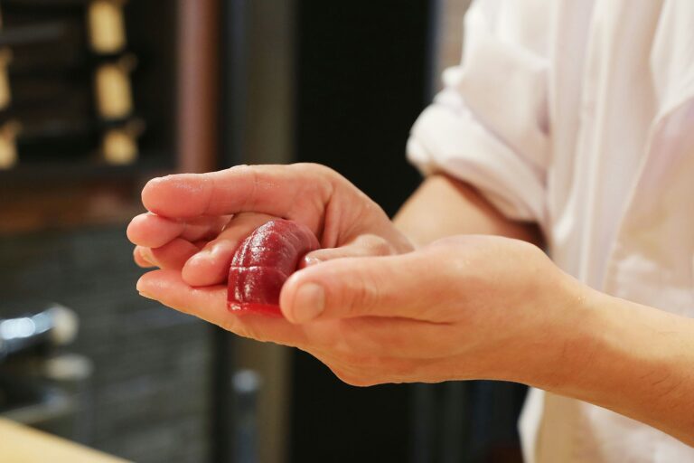 A sushi chef’s hands gently shaping rice and fresh fish behind the counter, capturing the intimate moment of sushi preparation.