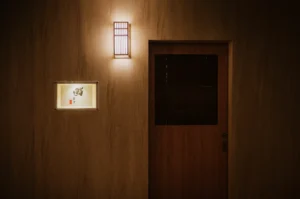Minimalist wooden door with a dark panel, illuminated by a wall-mounted lantern. Next to the door, a lit frame displays Japanese calligraphy.
