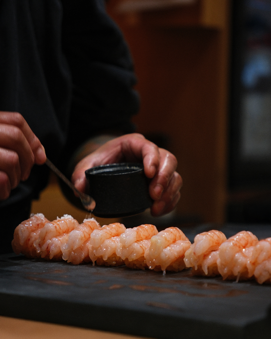 A close-up of Chef Masa delicately brushing seasoning onto sushi, highlighting the attention to balance, texture, and subtle flavour in each piece.