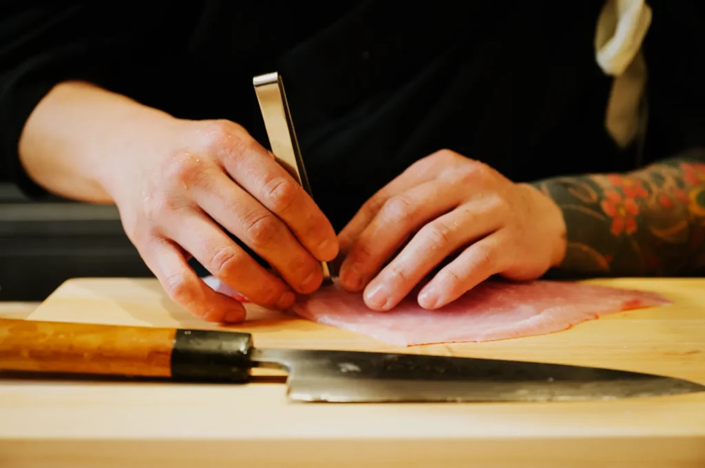 Chef Masa delicately trims a raw fish with tweezers on a wooden board, beside a sharp knife. The person’s arm shows detailed tattoos, conveying precision and focus.