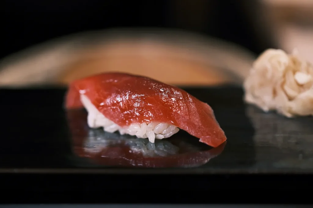 Close-up of tuna nigiri sushi on dark plate, showcasing glossy red fish atop white rice. Ginger garnish on the side; elegant and minimalist presentation.