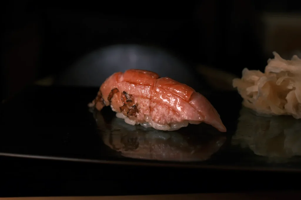 Close-up of a sushi piece with glossy marbled fish atop rice, served on a black plate. Soft lighting enhances its texture, paired with pale pickled ginger.