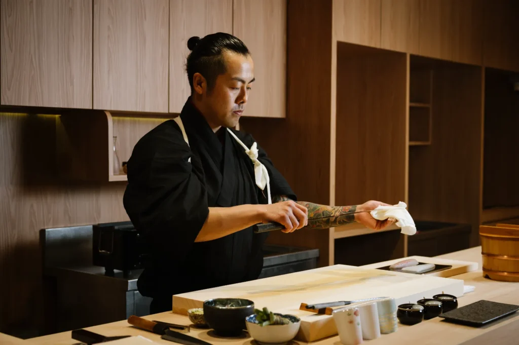 Chef Masa in a black kimono, with a focused expression, wipes a knife in a modern wooden kitchen. Ingredients and utensils are neatly arranged, creating a calm, professional atmosphere.