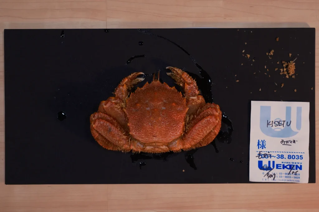 Cooked crab on a black tray with some breadcrumbs around it, accompanied by a business card with Japanese text, against a wooden table backdrop.
