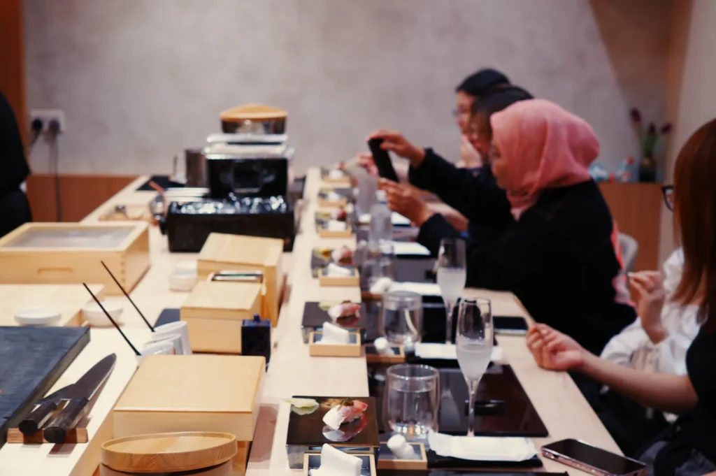 A group of people seated at a sushi bar, enjoying a meal. The atmosphere is casual and focused, with wooden trays and glasses on the counter.