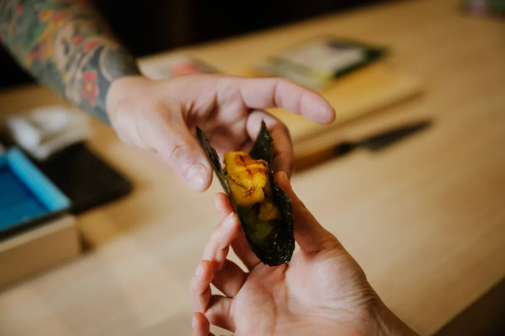 Close-up of chef Masa passing a seaweed-wrapped sushi to a customer's hand over a wooden table, conveying a sense of sharing and culinary craftsmanship.
