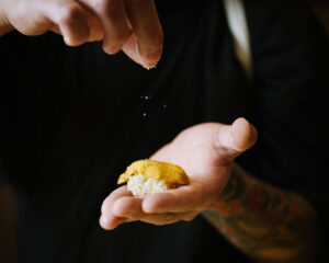 The chef’s hand gently sprinkles sea salt over freshly formed sushi, enhancing the natural sweetness of the fish under soft restaurant lighting.