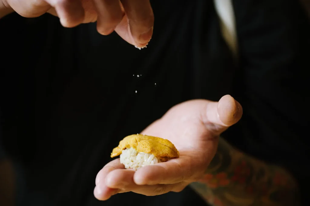 Chef Masa's hand delicately sprinkles salt over a piece of sushi topped with sea urchin, held in the other hand. The setting is intimate, focused on culinary craftsmanship.