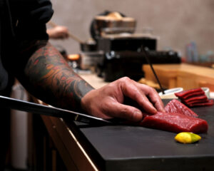 A close-up of Chef Masa’s hands carefully slicing fresh tuna with a sharp knife on a wooden board, highlighting the precision and craftsmanship behind each cut.