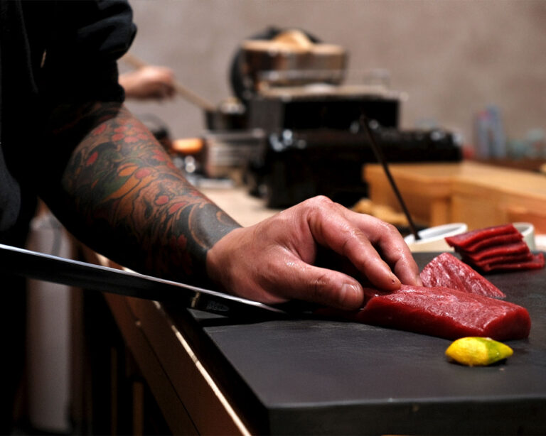 A close-up of Chef Masa’s hands carefully slicing fresh tuna with a sharp knife on a wooden board, highlighting the precision and craftsmanship behind each cut.