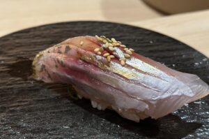 A piece of Iwashi (sardine) nigiri sushi topped with white sesame seeds and a sliver of green onion, served on a textured black stone plate.