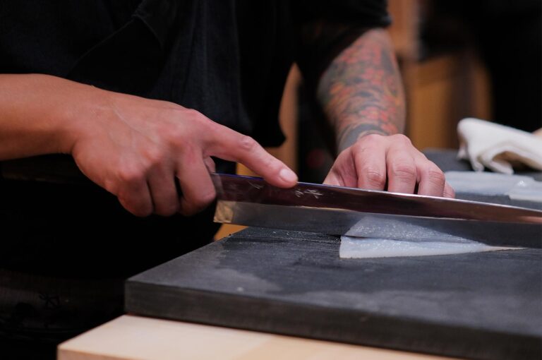 Close-up of a professional sushi chef with tattoos using a traditional Japanese yanagiba knife to thinly slice a piece of white fish on a dark cutting board.