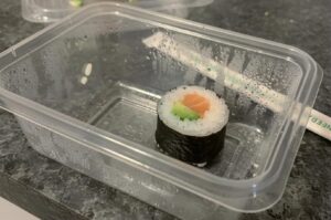 A single salmon and avocado maki sushi roll inside a clear plastic takeout container with chopsticks in the background.