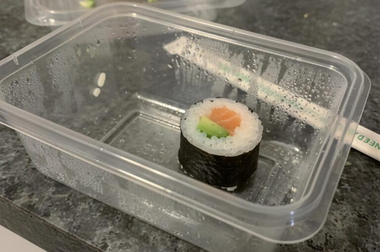 A single salmon and avocado maki sushi roll inside a clear plastic takeout container with chopsticks in the background.