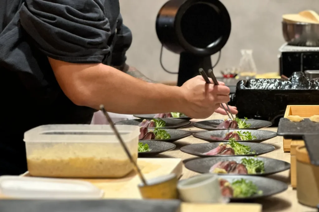 Chef Masa meticulously arranges garnishes on several charcoal plates with precision using chopsticks, preparing for a sophisticated dining presentation.