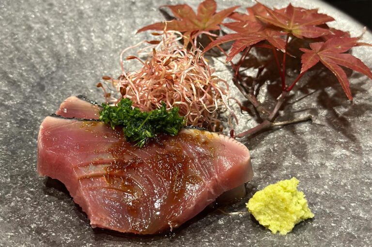 Two thick slices of seared Katsuo (bonito) sashimi garnished with green herbs and sprouts, served with a side of fresh wasabi and decorative red maple leaves.