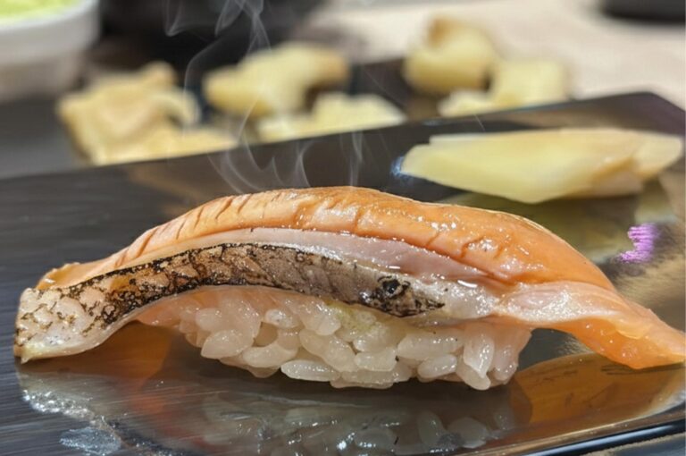 A piece of Aburi (torched) salmon nigiri sushi with a visible wisp of steam, featuring charred skin and a glossy glaze, served on a black plate.