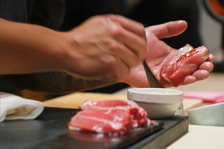 Action shot of a sushi chef's hands delicately brushing nikiri shoyu (soy sauce) onto a piece of fatty tuna nigiri held in their palm.