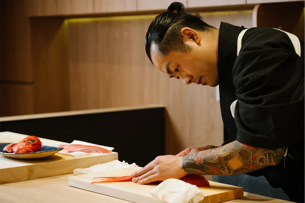 A focused Chef Masa with tattooed arms skillfully slices salmon on a wooden counter. A plate with fish head and fillets rests nearby, in a warm-lit kitchen.