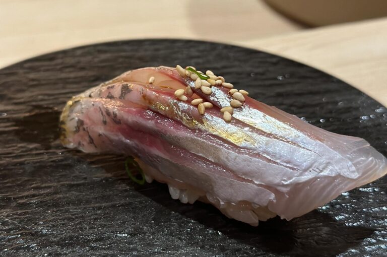 A close-up of a shimmering piece of Aji (horse mackerel) nigiri sushi, featuring silver skin and topped with sesame seeds on a dark plate.