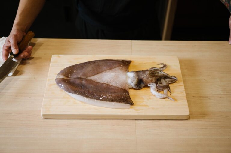 A large, fresh whole squid laid out on a light wood cutting board, with a chef’s hand holding a traditional Japanese knife to the left.
