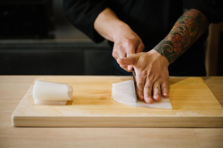 A professional sushi chef with tattoos using a Japanese knife to slice a clean, white raw squid mantle on a wooden board.