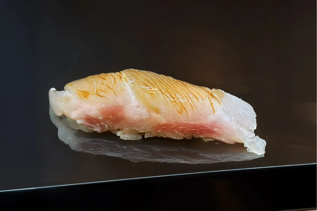 Close-up of a single sushi nigiri piece on a dark surface. The sushi features a delicate pink fish slice on top of white rice, conveying freshness and elegance.