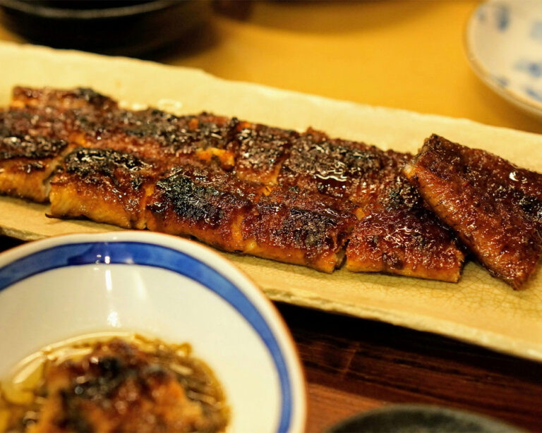 A serving of grilled eel presented on a simple plate, with the surface lightly charred and glazed, highlighting the smoky notes and the moist texture of the eel.