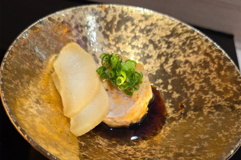 A circular slice of Ankimo (monkfish liver) topped with chopped scallions and served with pickled daikon radish in a hammered gold bowl with ponzu sauce.