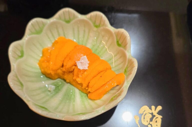 Bright orange, high-quality sea urchin (uni) tongues topped with a single flake of sea salt, served in a decorative green and white scalloped bowl.