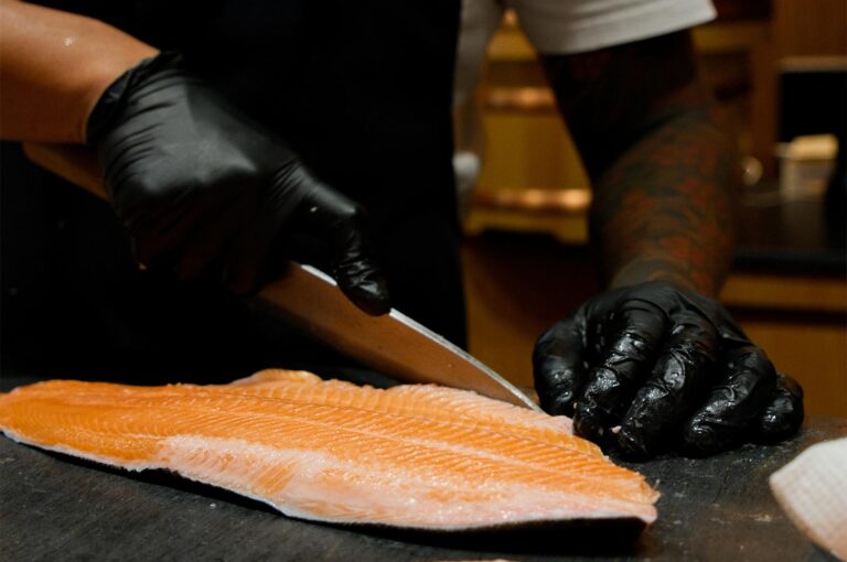 Professional sushi chef wearing black gloves using a sharp knife to carefully slice a fresh, marbled salmon fillet.