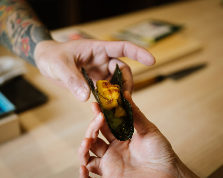 The chef’s hand presents a freshly prepared piece of sushi directly to the guest, creating an intimate omakase dining moment.