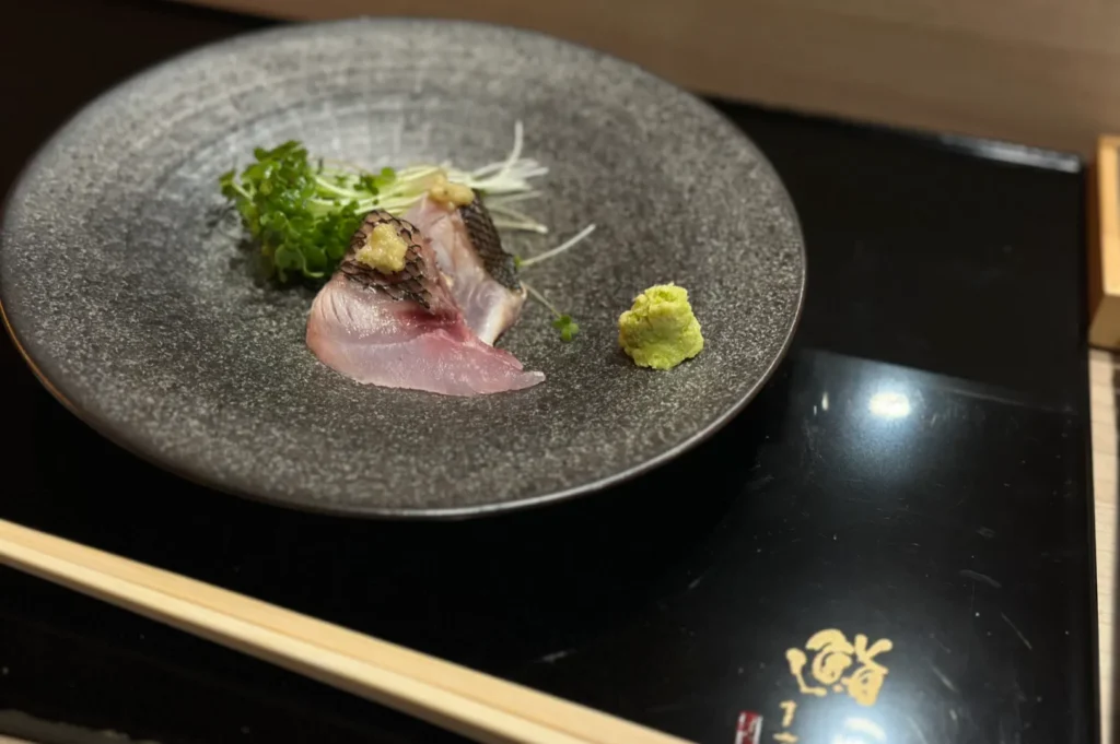 Slices of sashimi with garnish on a textured gray plate. Wasabi is placed beside the fish, creating an elegant and fresh presentation.