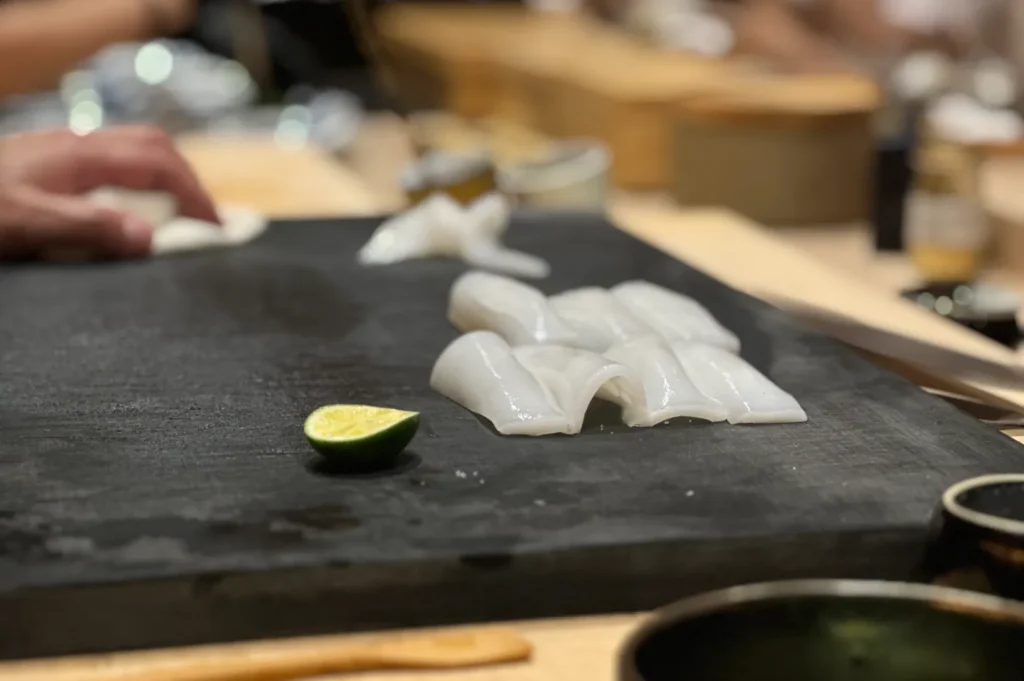 Sliced squid placed neatly on a dark cutting board beside a halved lime, with a hand holding a cloth in the blurred background, creating a fresh sushi preparation scene.