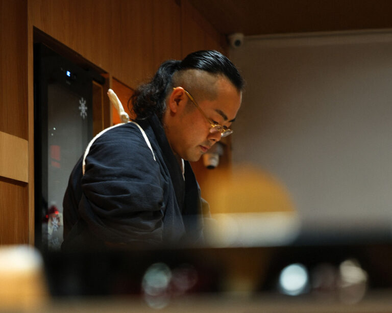 Chef Masa working silently behind the sushi counter, his calm movements and focused craftsmanship embodying years of discipline and devotion to omakase dining.