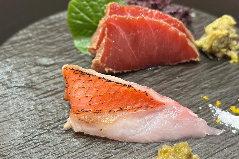 Artistic plating of seared tuna sashimi and white fish served with fresh wasabi, garnishes, and a green shiso leaf.