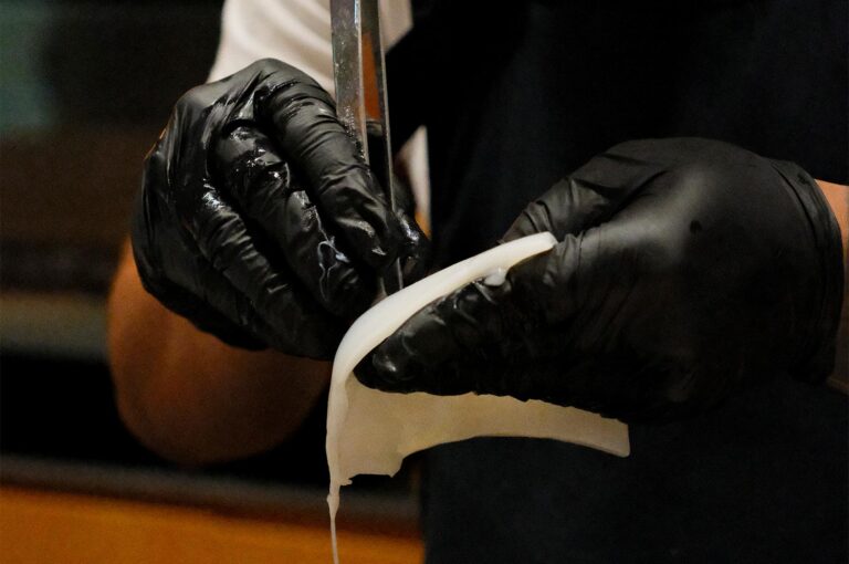 Close-up of a chef wearing black gloves using precision tongs to carefully clean and prepare a translucent piece of fresh squid (ika).