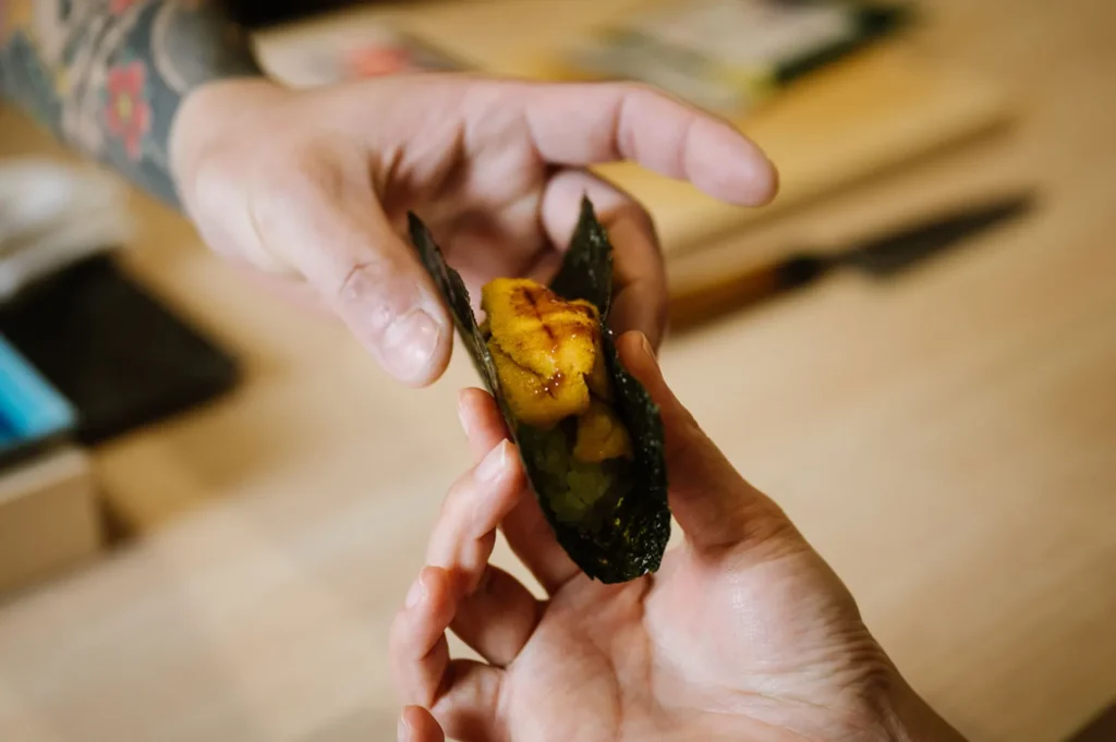 Hands exchanging a piece of sushi wrapped in seaweed, showcasing a vibrant yellow filling, set on a wooden table with a knife in the background.