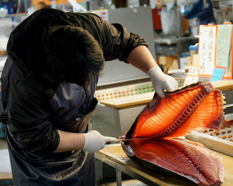 A fisherman cutting a large tuna at Toyosu Market in Japan, surrounded by market tools and worktables in a traditional wholesale setting.