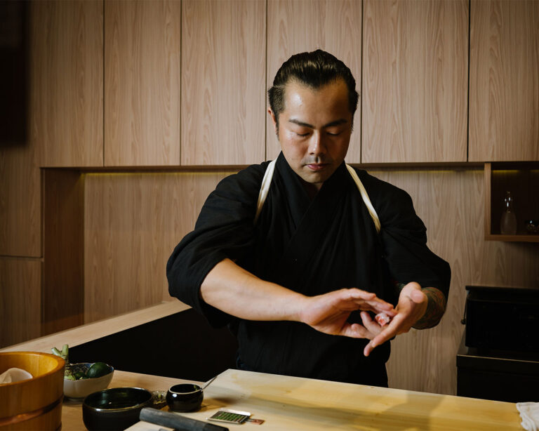 Chef Masa preparing a dish behind the sushi counter, focused on plating with careful hand movements and traditional technique.