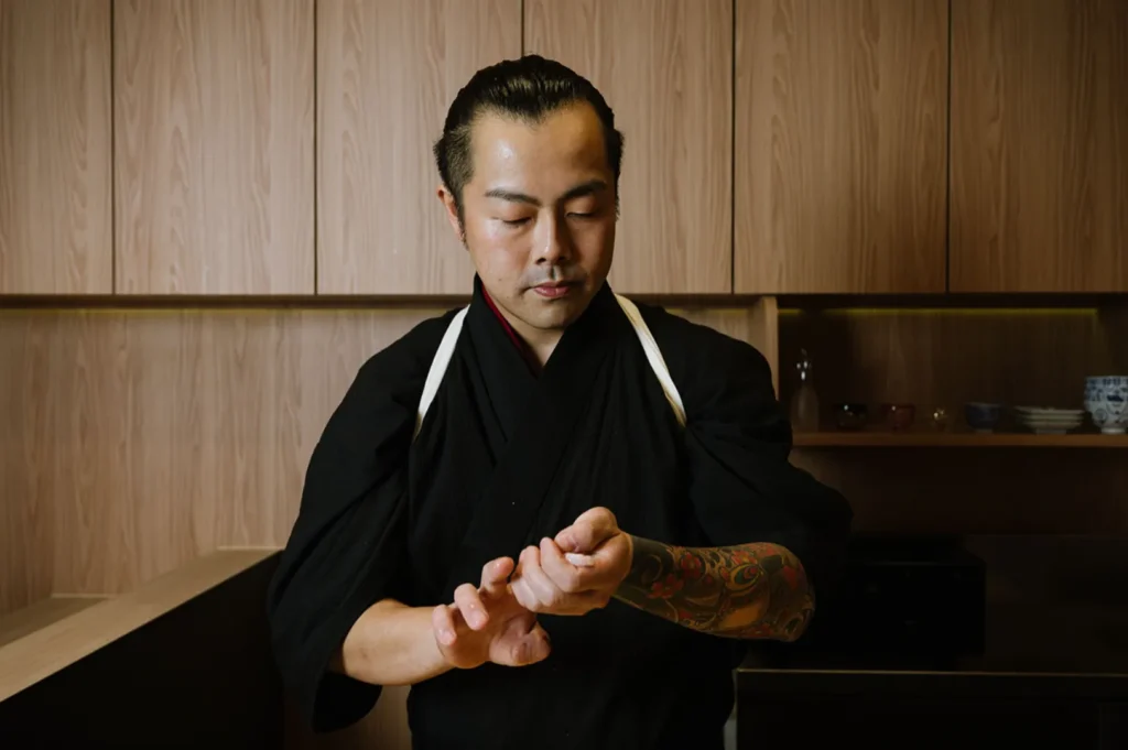 Chef Masa in a black uniform, with tattoos on his left arm, focuses intently as he shapes a sushi roll in a minimalist kitchen setting.