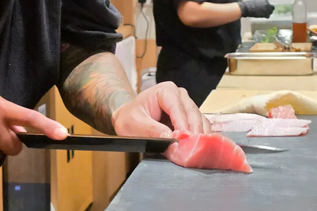 Professional sushi chef with tattoos slicing a fresh piece of raw tuna on a black cutting board in a Japanese restaurant.