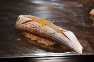 A piece of Kohada nigiri sushi with intricate decorative cuts and fresh yuzu zest garnish on a dark lacquer plate.