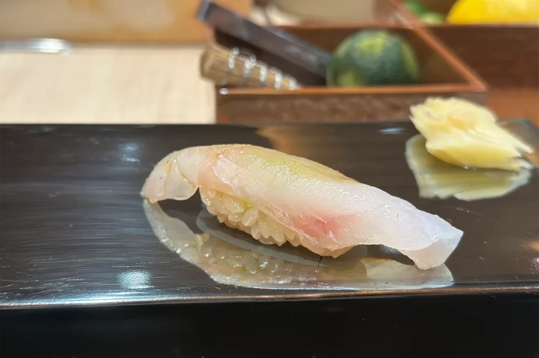 A piece of Madai (Red Sea Bream) nigiri sushi with translucent white fish on seasoned rice, served on a black lacquered plate with pickled ginger.