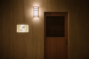 An elegant and minimalist wooden entrance to a Japanese sushi restaurant with a warm glowing wall lantern and a small calligraphic sign.