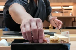 Close-up of a sushi chef's hand carefully placing a fresh piece of red snapper nigiri onto a black ceramic serving plate.