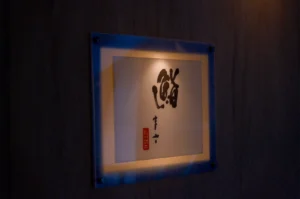 A minimalist, illuminated Japanese sushi restaurant sign featuring elegant black calligraphy on a white background, mounted on a dark wooden wall.