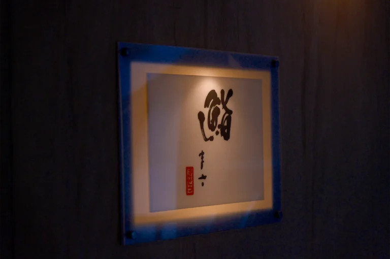 A minimalist, illuminated Japanese sushi restaurant sign featuring elegant black calligraphy on a white background, mounted on a dark wooden wall.